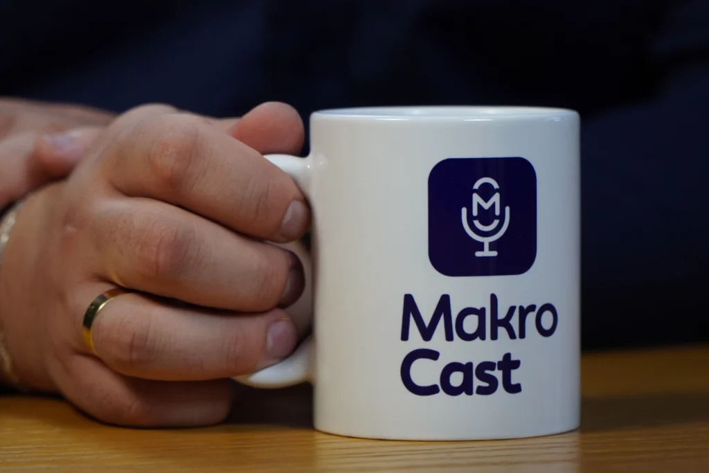 A imagem mostra uma mão segurando uma caneca com a inscrição 'MakroCast', promovendo o podcast para contadores.