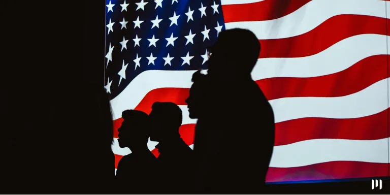 A imagem mostra a silhueta de pessoas aparentemente cantando o hino dos Estados Unidos e atrás a bandeira do país. A imagem representa o Mulher com cabelos loiros encaracolados usa um broche e segura uma bandeirinha dos Estados Unidos. A imagem representa o impacto das eleições estadunidenses no Brasil