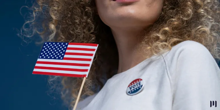 Mulher com cabelos loiros encaracolados usa um broche e segura uma bandeirinha dos Estados Unidos. A imagem representa o impacto das eleições estadunidenses no Brasil