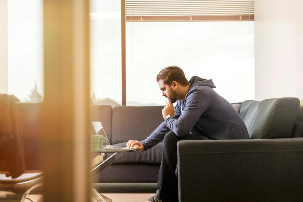 Homem sentado no sofá olha para a tela do computador onde está vendo a notícia sobre como ficará o Simples na Reforma Tributária