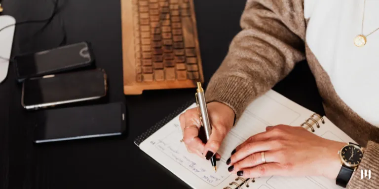 A imagem mostra a mão de uma mulher, escrevendo em uma agenda em cima da mesa de escritório.