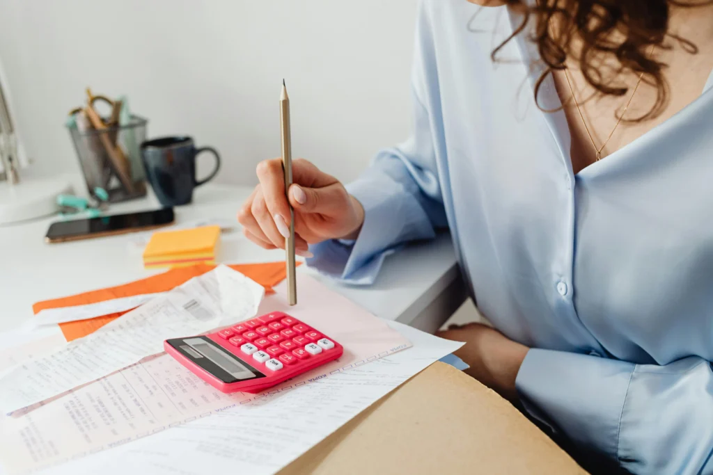 Mulher preocupada com as contas. A imagem foca em uma calculadora rosa em cima da mesa e a mulher aparece pela metade sem mostrar o rosto. A imagem ilustra a reportagem sobre Débito automático Receita Federal