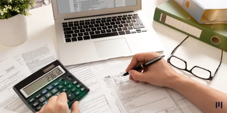 A imagem mostra uma pessoa sentada a uma mesa, com um notebook, uma calculadora a sua frente, preenchendo um papel com uma caneta.