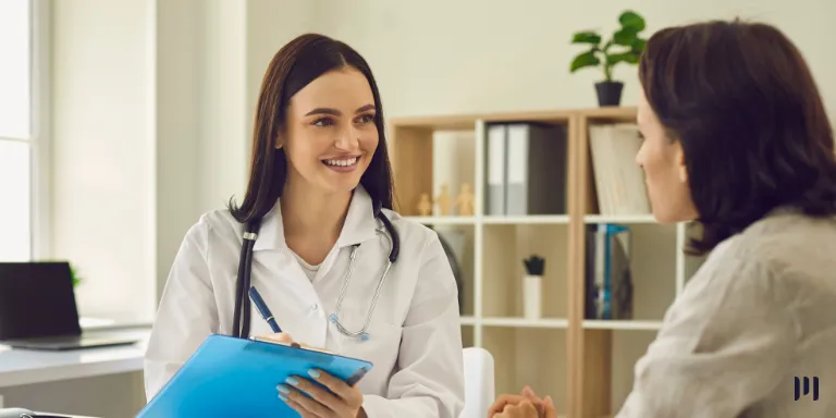 A imagem mostra uma médica vestida em seu jaleco e segurando uma prancheta azul, conversando com uma paciente em seu escritório.