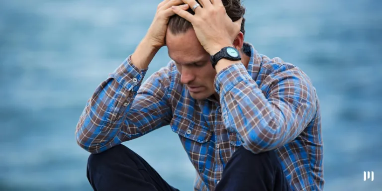 Homem está triste sentado no chão com o fundo de um lago embaçado. Ele está com a camisa xadrez e coloca as mãos na cabeça em sinal de desespero pois está com o nome sujo.