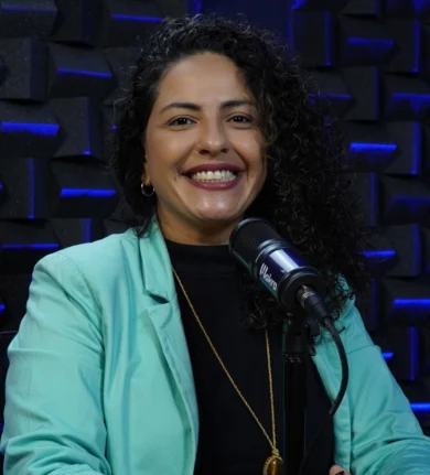 Flávia Rodrigues sorrindo, com cabelo cacheado e usando blusa preta com blazer ciano, sentada em uma mesa com um microfone à sua frente. Fundo com painéis acústicos.