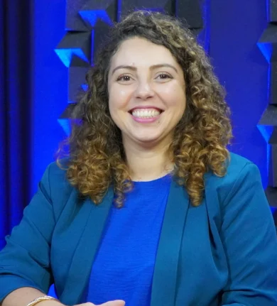 Vanessa Oliveira, sorrindo, cabelos cacheados, vestindo blusa azul e blazer da mesma cor, está sentada em uma mesa com painéis acústicos ao fundo.